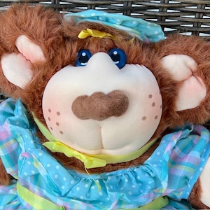 Op de afbeelding: Een pluche teddybeer met bruine vacht, blauwe ogen en een lichtbruine neus. De beer draagt een blauw-witte stippenmuts en een blauw geruite jurk met gele accenten. De beer heeft roze wangen en een bruine mond.
