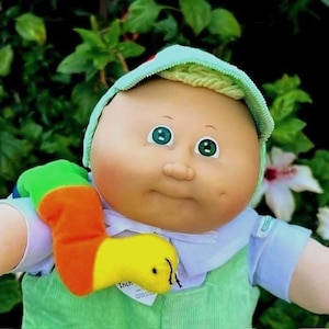 May include: A vintage Cabbage Patch Kid doll with a light green hat and overalls. The doll has green eyes, rosy cheeks, and a soft, plush appearance. A colorful worm-like toy is attached to the doll's shoulder.