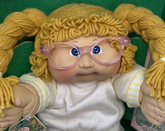 May include: A Cabbage Patch Kids doll with blonde yarn pigtails and pink-rimmed glasses. The doll has blue eyes and is wearing a white shirt with yellow stripes. A tag with the Cabbage Patch Kids logo is visible.