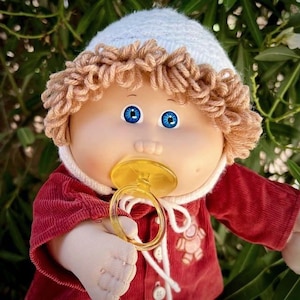Op de afbeelding: Een vintage Cabbage Patch Kid pop met blauwe ogen, met een witte gebreide muts, een rode corduroy jas en een gele fopspeen. De pop heeft garen haar en een witte slab. De jas heeft een teddybeer ontwerp.