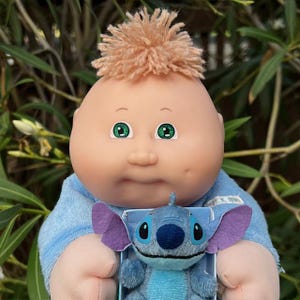 May include: A Cabbage Patch Kids doll with green eyes and light brown hair, wearing a blue shirt, holding a small blue Stitch plush toy with purple ears. The plush toy is in a small box.