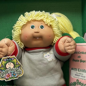 Może przedstawiać: Lalka Cabbage Patch Kids z blond włosami z przędzy i zielonymi oczami. Lalka ma na sobie szary sweter z czerwonymi wykończeniami i białą grafiką kota. Etykieta głosi "Cabbage Patch Kids". Widoczny jest różowy akt urodzenia.