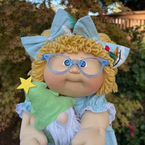 May include: A vintage doll with blonde curly hair, blue glasses, and a light blue dress. The doll is holding a green felt Christmas tree with a yellow star and a gnome. A light blue bow is in the doll's hair.