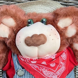 以下が含まれることがあります： 麦わら帽子、赤いバンダナ、青いオーバーオールを着たぬいぐるみ。クマは茶色の毛皮、そばかすのある薄いピンク色の顔、緑色の目をしています。バンダナは白いペイズリー柄です。