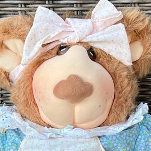 Op de afbeelding: Een pluchen teddybeer met een lichtbruin gezicht, bruine vacht en een roze strik met een bloemenpatroon. De beer draagt een lichtblauwe bloemenjurk met een witte kanten kraag. De neus van de beer is bruin en zijn ogen zijn donker.