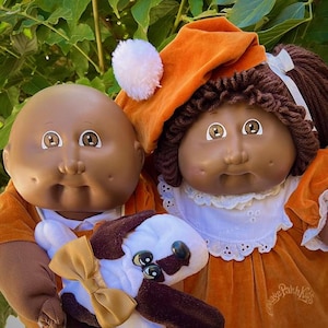 Puede incluir: Dos muñecas Cabbage Patch Kids con piel morena y ojos marrones. Una muñeca lleva un conjunto y un sombrero naranja con un pompón blanco. La otra muñeca lleva un vestido y un sombrero naranja. Un juguete de peluche de perro con un lazo está en primer plano.