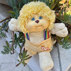 May include: A plush doll with light skin, bright blue eyes, and a small brown nose. The doll has textured yellow yarn hair and wears light yellow corduroy overalls with rainbow-striped straps and an embroidered orange and white unicorn graphic.