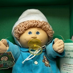 Könnte beinhalten: Cabbage Patch Kids Puppe mit blauem Cord-Outfit, weißer Strickmütze und gelbem Schnuller. Die Puppe hat hellbraunes lockiges Haar und blaue Augen. Ein Etikett mit dem Cabbage Patch Kids-Logo ist angebracht. Die Puppe befindet sich in der Originalverpackung.