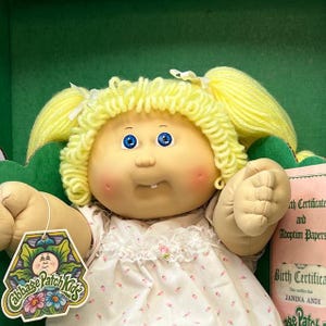 May include: A Cabbage Patch Kids doll with blonde yarn hair in pigtails. The doll has blue eyes, rosy cheeks, and a single tooth. It wears a white dress with a floral pattern and lace trim. A tag with the Cabbage Patch Kids logo is attached.