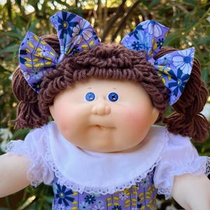 May include: A vintage doll with brown yarn hair styled in pigtails and a matching yarn fringe. The doll has blue eyes, rosy cheeks, and a white dress with a purple floral print. The doll is wearing two large purple bows with a floral pattern.