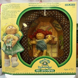Puede incluir: Un juego de muñecas vintage Cabbage Patch Kids Pin-Ups Minis en su embalaje original. La caja presenta dos muñecas, una que sostiene una muñeca más pequeña y otra dentro de una casa club en miniatura. El embalaje incluye texto en inglés y francés.