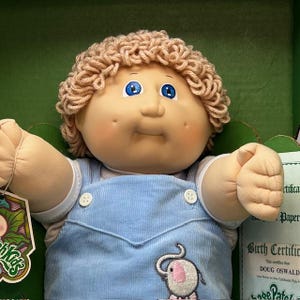 May include: A Cabbage Patch Kids doll with a light skin tone, blue eyes, and light brown curly hair. The doll wears a blue corduroy overall with an embroidered elephant design. A tag with the Cabbage Patch Kids logo is visible.