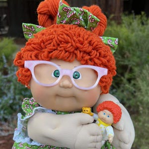 Könnte beinhalten: Eine Cabbage Patch Kid-Puppe mit orangefarbenem Garnhaar, einer rosa Brille und einer grünen Schleife mit Zuckerstangen-Akzenten. Die Puppe hält eine kleinere Puppe und einen gelben Teddybären.