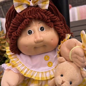 May include: A doll with brown yarn hair, a yellow and white gingham bow, and freckles on its cheeks. It wears a white and yellow gingham dress with two yellow buttons and a ruffled collar, while holding a small, light brown plush teddy bear.