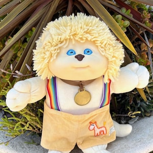 Puede incluir: Una muñeca de peluche vintage con una cara color crema, ojos azules y cabello rubio rizado. Lleva un collar marrón con un colgante dorado, pantalones cortos de pana y tirantes arcoíris. Un caballo de dibujos animados está bordado en los pantalones cortos.