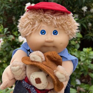 Könnte beinhalten: Cabbage Patch Kid-Puppe mit blondem, lockigem Haar und blauen Augen, trägt eine rote Kappe und ein blaues kariertes Hemd. Die Puppe hält einen Teddybären mit braunem Cowboyhut.
