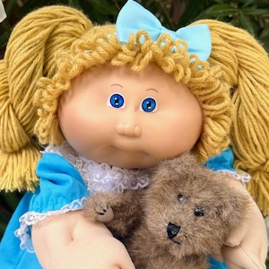 Puede incluir: Una muñeca vintage con coletas rubias de hilo, ojos azules y un lazo azul claro. La muñeca viste un vestido azul con cuello de encaje blanco y sostiene un pequeño osito de peluche marrón.