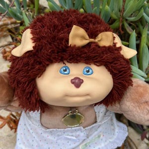 Puede incluir: Una muñeca vintage de Cabbage Patch Kids con un peinado de felpa marrón rojizo y un lazo color canela. La muñeca tiene ojos azules, una nariz pequeña y un vestido blanco con pequeños lunares azules. Un colgante dorado cuelga de su cuello.