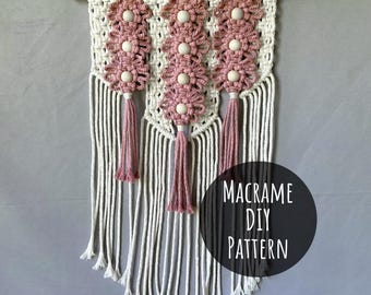 Peut inclure: Suspension murale en macramé avec une baguette en bois naturel. Le motif présente trois rangées de motifs floraux roses avec des perles en bois, des glands et des franges blanches. Le texte "Macramé DIY Pattern" est dans un cercle noir.