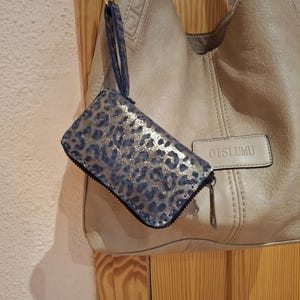 May include: A blue and gold leopard print wallet with a wrist strap hangs next to a beige handbag. The wallet has a zipper closure and a black trim. The handbag has a gold-colored handle and the brand name "OTSLUMU" is visible.