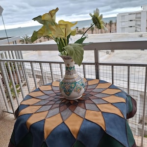 May include: A round table with a patchwork leather tablecloth in blue, green, brown, and tan. A decorative vase with a floral design holds yellow flowers. The scene is set on a balcony overlooking the sea.
