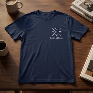 Könnte beinhalten: Marineblaues T-Shirt mit weißem Grafikdesign auf der Brust. Das Design enthält die Buchstaben "M" auf beiden Seiten eines Herzens und eines Kreises, mit dem Text "Daniel & Hanna" darunter. Das Shirt wird auf einer Holzoberfläche präsentiert.