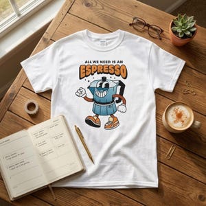 Può includere: Una t-shirt bianca a maniche corte con la grafica di una caffettiera moka blu raffigurata come un personaggio con braccia, gambe e scarpe da ginnastica. Sopra il personaggio, il testo "ALL WE NEED IS AN ESPRESSO" in arancione con bordi neri.