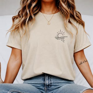 Könnte beinhalten: Beiges T-Shirt mit einer schwarzen Strichzeichnung einer Sonne über Wasser auf der linken Brust. Das Shirt hat kurze Ärmel und eine lockere Passform. Die Person trägt eine blaue Jeans und hält Schuhe.