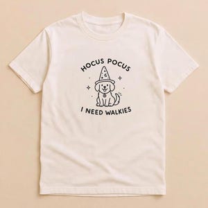 Könnte beinhalten: Ein cremefarbenes Kurzarm-T-Shirt mit einer schwarzen Strichzeichnung eines Welpen, der einen Zaubererhut trägt. Der Text über dem Welpen lautet HOCUS POCUS und der Text darunter I NEED WALKIES, ergänzt durch kleine Sternsymbole.