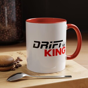May include: White ceramic mug with a red interior and handle. The mug features the text "DRIFT KING" in black and red, with a small crown graphic. The mug is on a wooden surface with a spoon and a pastry.