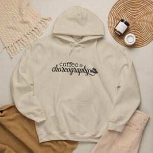 May include: A sand-colored hooded sweatshirt featuring a front kangaroo pocket and long sleeves. The chest displays the black text "coffee & choreography" accompanied by a small graphic of two dance shoes positioned to the right of the lettering.