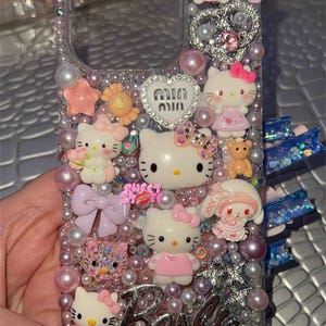 May include: A clear phone case decorated with Hello Kitty characters, pearls, glitter, and rhinestones. The case has a pink, white, and silver color scheme and says "Barbie" on the bottom.