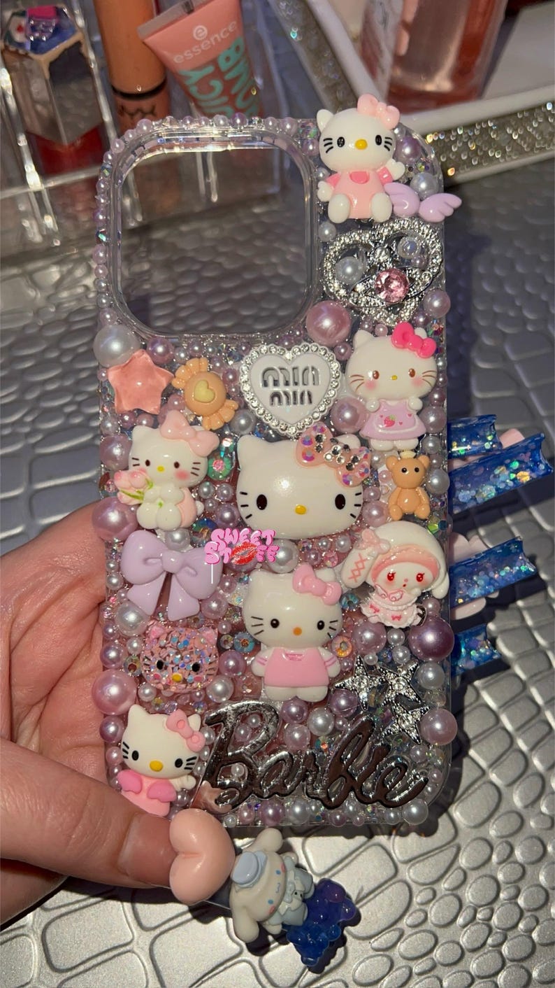 May include: A clear phone case decorated with Hello Kitty characters, pearls, glitter, and rhinestones. The case has a pink, white, and silver color scheme and says "Barbie" on the bottom.