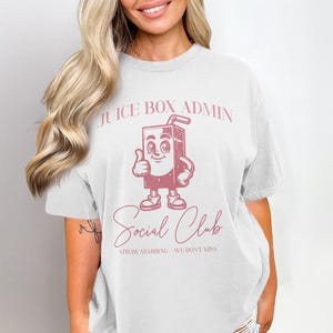 May include: White t-shirt featuring a pink juice box character with a thumbs up. The text "JUICE BOX ADMIN" is above the character, and "Social Club" is below. The shirt also has the text "STRAW STABBING WE DON'T MISS".