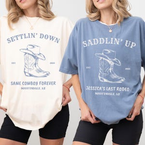 May include: Two oversized t-shirts, one cream and one blue, featuring a graphic of a cowboy hat and boots. The cream shirt reads SETTLIN' DOWN, SAME COWBOY FOREVER, SCOTTSDALE, AZ. The blue shirt reads SADDLIN' UP, JESSICA'S LAST RODEO, SCOTTSDALE, AZ.