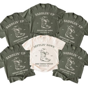 May include: Five olive green t-shirts and one cream t-shirt featuring a graphic of a cowboy hat and boot. The olive shirts read SADDLIN' UP, JESSICA'S LAST RODEO, SCOTTSDALE, AZ. The cream shirt reads SETTLIN' DOWN, SAME COWBOY FOREVER, SCOTTSDALE, AZ.