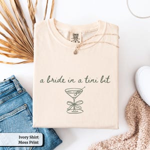 May include: An ivory-coloured t-shirt with the text "a bride in a tini bit" and a moss green martini glass illustration. A gold necklace and blue jeans are visible. The shirt is folded.