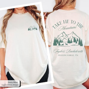 May include: Ivory-colored t-shirt with a light green mountain graphic on the front and back. The back of the shirt features the text "TAKE ME TO THE Mountains" above a mountain and tree design, with "Taylor's Bachelorette" and "PIGEON FORGE, TN" below.