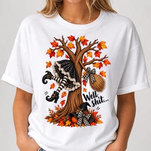 May include: A white short-sleeve t-shirt featuring a graphic of a tree with autumn leaves, a witch's legs in striped stockings and black boots, a broom, a pumpkin, and a black hat. The text "Well shit..." is printed in black script.