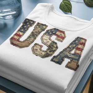 May include: A white t-shirt featuring the letters USA in a large, distressed applique font. The letters are filled with a patchwork design of the American flag, camouflage patterns, and denim textures with frayed edges and visible stitching.