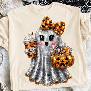 Pode incluir: Uma t-shirt cor de creme com um gráfico de um fantasma com textura de bola de discoteca prateada e um laço laranja. O fantasma segura um copo de café com a palavra BOO e um cesto em forma de abóbora cheio de doces coloridos.