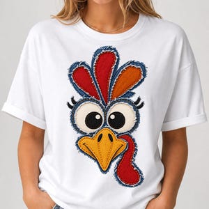 Puede incluir: Una camiseta blanca de manga corta con un gran gráfico colorido de una cara de pavo. El diseño utiliza telas superpuestas con bordes deshilachados, mostrando un pico amarillo, una carúncula roja y plumas rojas y naranjas vibrantes.