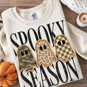May include: A cream-colored crewneck sweatshirt featuring the black text SPOOKY SEASON. The design includes three ghost-shaped patches with distinct patterns: one with green camouflage, one with a tan leopard print, and one with a tan checkerboard.