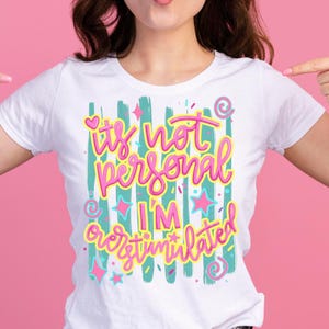 Puede incluir: Una camiseta blanca de manga corta con un estampado gráfico de pinceladas verticales en color verde azulado y el texto "it's not personal I'M overstimulated" en letras cursivas de color rosa brillante y amarillo, con pequeñas estrellas, corazones y espirales rosas.