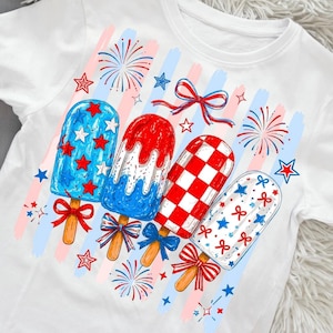 May include: A white t-shirt featuring a graphic print of four popsicle treats in red, white, and blue. The design includes star patterns, a red and white checkered pattern, and red and blue bows on the wooden sticks, accented by firework illustrations.