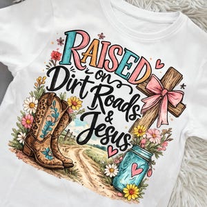 Puede incluir: Una camiseta blanca con un estampado gráfico que incluye el texto "Raised on Dirt Roads & Jesus". El diseño muestra un par de botas vaqueras marrones con estampado de leopardo, una cruz de madera con un lazo rosa, un frasco azul y flores silvestres.