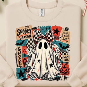 Puede incluir: Una sudadera beige de cuello redondo con un estampado gráfico de un fantasma blanco con un lazo a cuadros blanco y negro. El diseño incluye los textos "SPOOKY SEASON", "just a little boo!" y "GHOST", junto con murciélagos, estrellas y una calabaza.