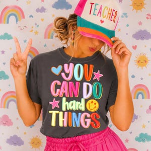 May include: A charcoal gray t-shirt featuring the colorful text "YOU CAN DO hard THINGS" with a heart, stars, and a smiley face. A trucker hat with a white front panel, the word "TEACHER" in rainbow letters, and a pink mesh back is worn.