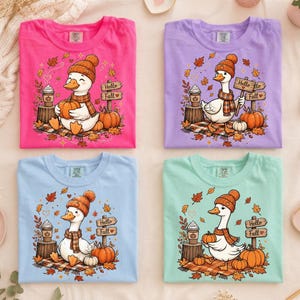 May include: Four t-shirts in bright pink, light purple, light blue, and mint green. Each features a graphic of a white goose wearing an orange knit hat and plaid scarf, holding a pumpkin, with a sign that reads Hello Fall and a coffee cup.