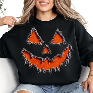 May include: A black long-sleeve sweatshirt featuring a large, central jack-o'-lantern face graphic. The eyes, nose, and wide, jagged mouth are filled with a textured orange fabric, outlined by frayed, distressed edges for a raw appearance.